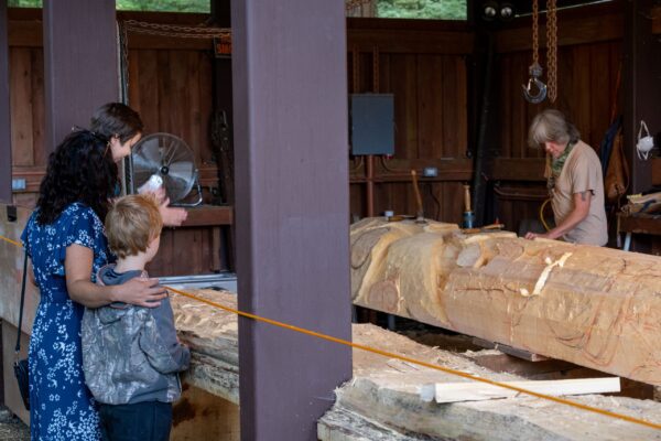 Family watching Tommy Joseph carve a totem pole in the carving shed at Sitka National Historical Park.
